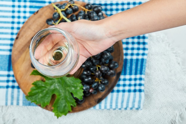 Un grappolo di uva nera sul piatto di legno con foglia mentre la mano tiene un bicchiere vuoto