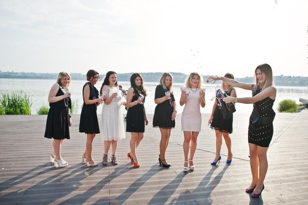Un gruppo di 8 ragazze indossano il nero e 2 spose alla festa di addio al nubilato contro la spiaggia soleggiata che beve champagne
