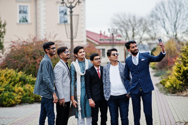 Un gruppo di sei uomini indiani del sud asiatico nel tradizionale abbigliamento casual e da lavoro in piedi e facendo selfie insieme al telefono cellulare
