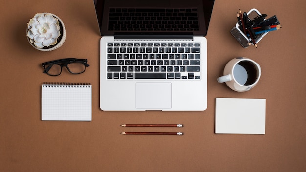 Un laptop aperto; con tazza di caffè; carta; matite; occhiali e blocco note su sfondo di carta marrone
