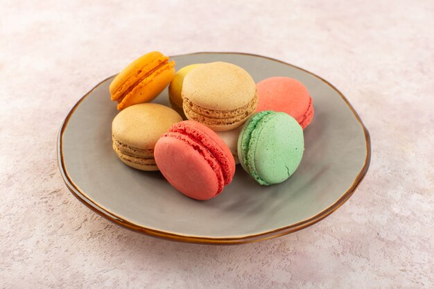 Un macarons francesi variopinti di vista frontale all'interno del piatto sulla torta dolce dello zucchero del biscotto dello scrittorio rosa