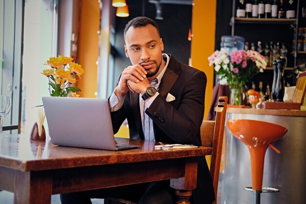 Un maschio americano nero premuroso vestito con un vestito elegante usando un computer portatile in un caffè.