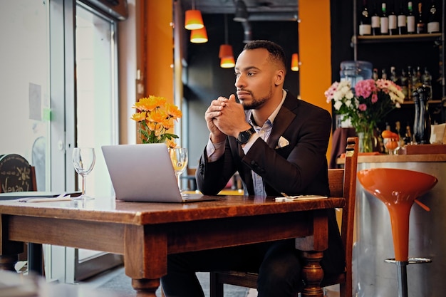 Un maschio americano nero premuroso vestito con un vestito elegante usando un computer portatile in un caffè.