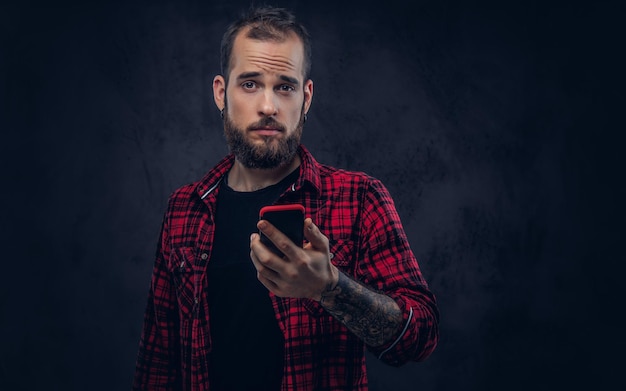 Un maschio barbuto hipster con un tatuaggio sulle braccia che tiene il telefono, guardando una macchina fotografica
