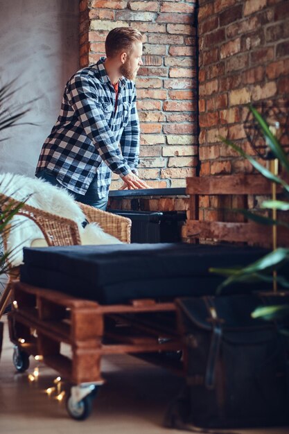 Un maschio biondo barbuto hipster vestito con jeans e camicia in pile in posa in una stanza con interni soppalcati.