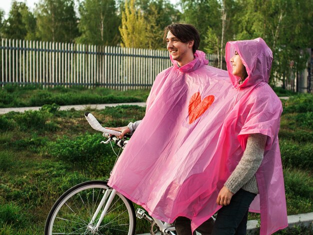 Un maschio e una femmina in un impermeabile di plastica rosa condiviso che camminano con una bicicletta per un appuntamento