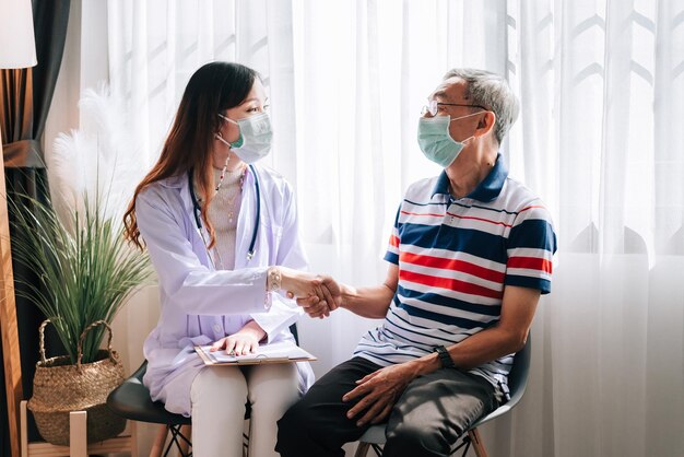 Un medico asiatico visita ed esamina un uomo anziano a casa che si stringe la mano dopo aver terminato il controllo sanitario, i servizi ospedalieri e indossando una maschera per proteggere il covid19