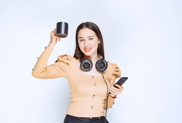 Un modello attraente della donna che tiene una tazza con il telefono cellulare e le cuffie.