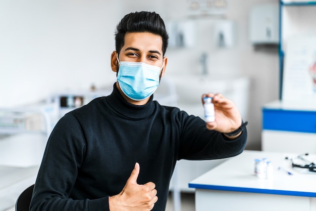 Un moderno uomo indiano tiene in mano un vaccino e sorride, indossando una maschera protettiva.