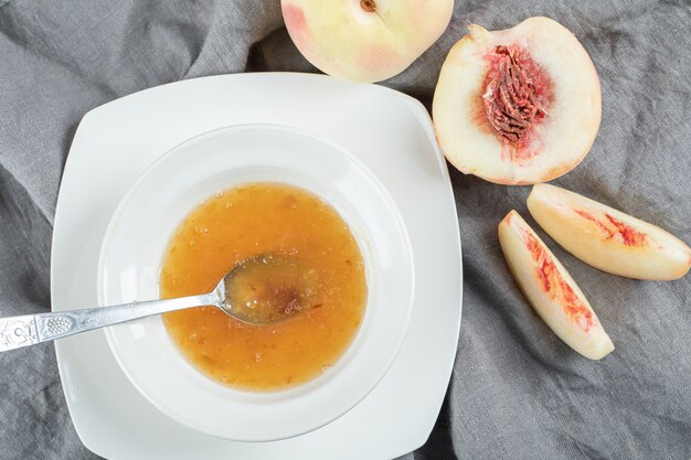 Un piatto bianco con marmellata e pesche sulla tovaglia grigia.