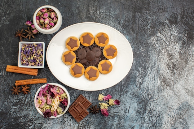 Un piatto di biscotti assortiti con diverse ciotole di fiori secchi e barretta di cioccolato su fondo grigio