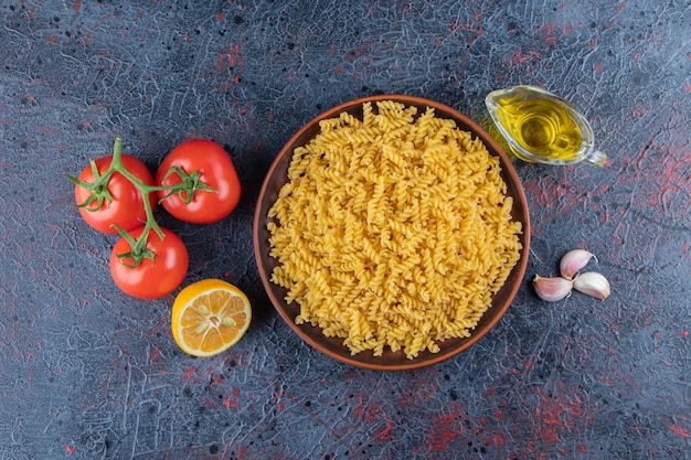Un piatto di pasta a spirale cruda con olio e pomodori rossi freschi su una superficie scura.
