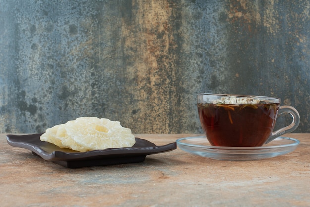 Un piatto scuro pieno di ananas sano essiccato e tazza di tè