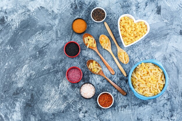 Un po 'di pasta cruda con spezie in ciotole e cucchiai di legno su sfondo grigio intonaco, vista dall'alto.