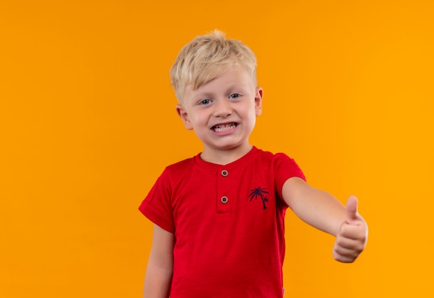 Un ragazzino sveglio con i capelli biondi e gli occhi azzurri che indossa la maglietta rossa che mostra i pollici in su mentre osserva su una parete gialla