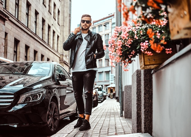 Un ragazzo alla moda con occhiali da sole vestito con una giacca di pelle nera e jeans parla da uno smartphone mentre si trova vicino a un'auto di lusso in una strada della vecchia Europa.