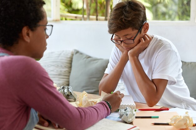 Un ragazzo asiatico serio e concentrato tiene le mani sulle guance, indossa occhiali trasparenti, si concentra sul blocco note, ascolta le spiegazioni del tutor, si siede l'uno di fronte all'altro in un caffè con terrazza, risolve compiti problematici