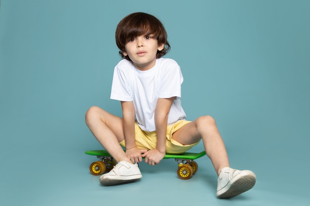 Un ragazzo carino vista frontale in maglietta bianca e pantaloncini gialli in sella a skateboard verde sullo spazio blu
