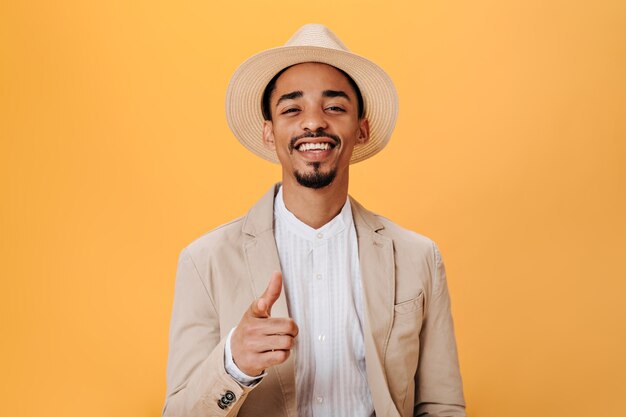 Un ragazzo con un cappello alla moda e una giacca beige sorride e mostra il dito alla telecamera