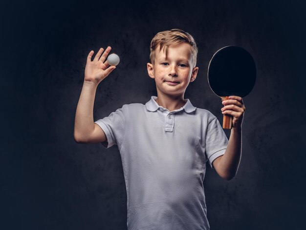 Un ragazzo dai capelli rossi carino vestito con una maglietta bianca tiene una racchetta da ping-pong e una palla in uno studio. Isolato su uno sfondo scuro con texture.