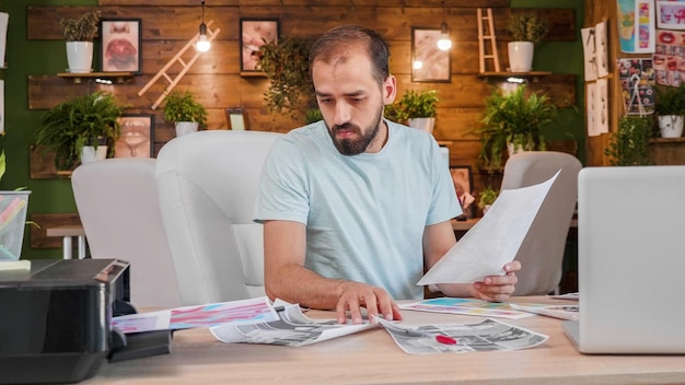 Un ragazzo di design seduto al tavolo e guardando alcune immagini stampate. Ufficio creativo