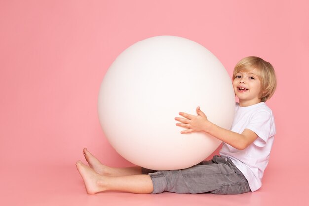 Un ragazzo felice biondo di vista frontale che gioca con la palla bianca in maglietta bianca sul pavimento rosa