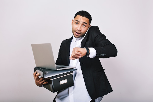 Un ragazzo impegnato e laborioso in camicia bianca e giacca nera parla al telefono, tiene in mano cartelle, laptop, guarda stupito all'orologio. Impiegato, tecnologia moderna, carriera.