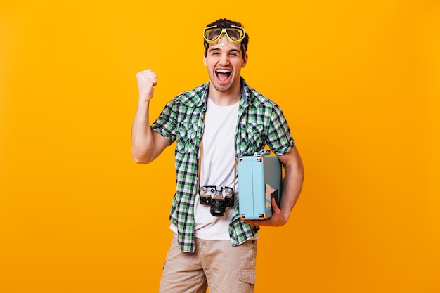 Un ragazzo in camicia verde e pantaloncini beige si rallegra emotivamente e stringe il pugno. L'uomo con la maschera subacquea, la fotocamera retrò e la valigia ride sullo spazio arancione.