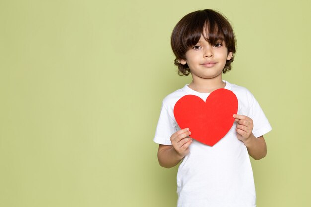 Un ragazzo sorridente del bambino di vista frontale in maglietta bianca che tiene forma del cuore sullo spazio colorato pietra