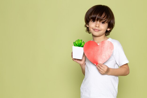 Un ragazzo sorridente del bambino di vista frontale in maglietta bianca che tiene forma del cuore sullo spazio colorato pietra