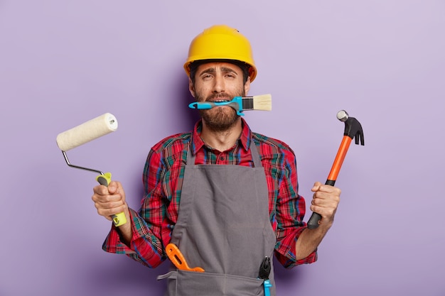 Un riparatore impegnato tiene strumenti da costruzione, fa riparazioni a casa, indossa elmetto giallo, grembiule, stand al coperto.