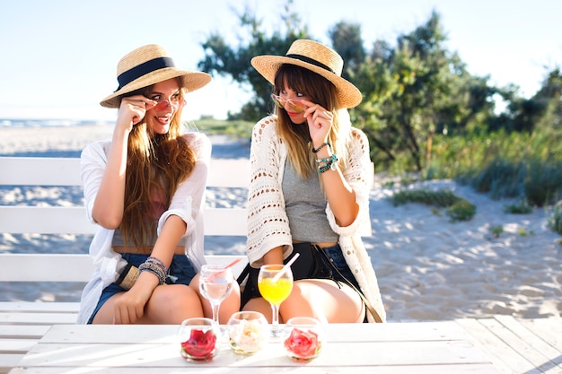 Un ritratto all'aperto di fanny di due sorelle ha battuto la ragazza degli amici divertendosi abbracci sorridendo e facendo smorfie sul bar sulla spiaggia, vestiti boho hipster, bevendo gustosi cocktail, vacanze estive sull'oceano.