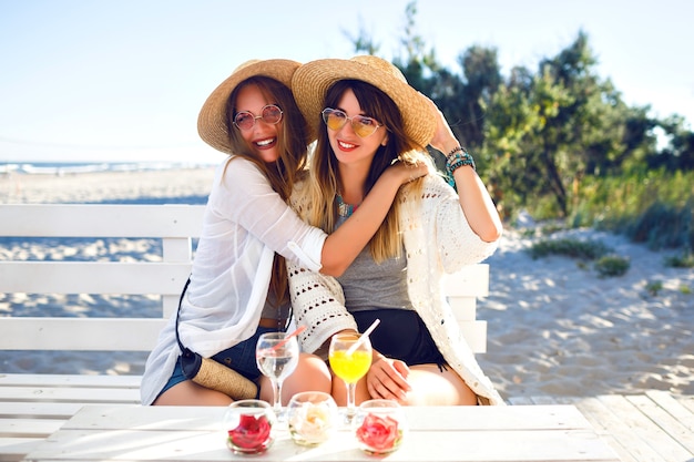 Un ritratto all'aperto di fanny di due sorelle ha battuto la ragazza degli amici divertendosi abbracci sorridendo e facendo smorfie sul bar sulla spiaggia, vestiti boho hipster, bevendo gustosi cocktail, vacanze estive sull'oceano.