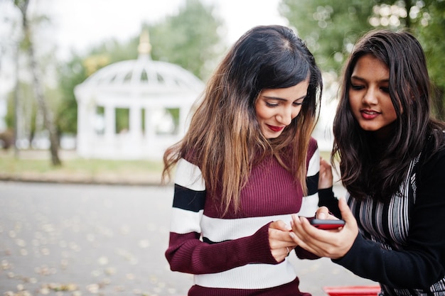 Un ritratto di due giovani belle ragazze adolescenti indiane o asiatiche del sud in vestito che si siedono sulla panchina e usano il telefono cellulare