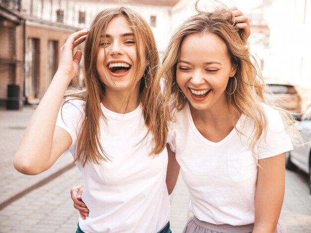 Un ritratto di due giovani belle ragazze sorridenti bionde dei pantaloni a vita bassa in vestiti bianchi della maglietta dell'estate d'avanguardia. . Divertirsi con i modelli positivi