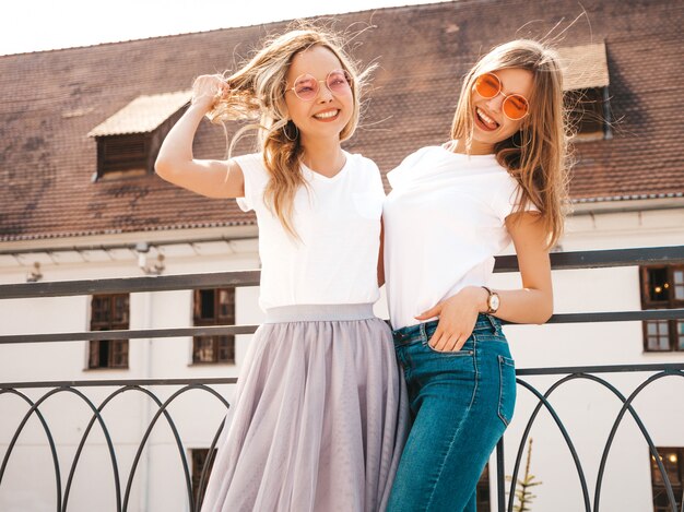 Un ritratto di due giovani belle ragazze sorridenti bionde dei pantaloni a vita bassa in vestiti bianchi della maglietta dell'estate d'avanguardia. . Divertirsi con i modelli positivi
