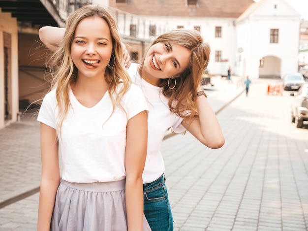 Un ritratto di due giovani belle ragazze sorridenti bionde dei pantaloni a vita bassa in vestiti bianchi della maglietta dell'estate d'avanguardia. . I modelli positivi mostrano la lingua