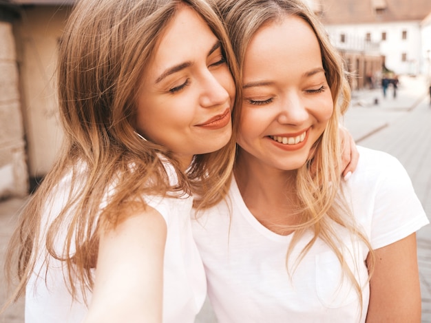 Un ritratto di due giovani belle ragazze sorridenti bionde dei pantaloni a vita bassa in vestiti bianchi della maglietta dell'estate d'avanguardia. . Modelli positivi che si divertono in occhiali da sole