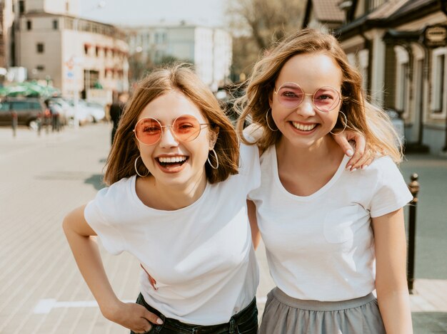 Un ritratto di due giovani belle ragazze sorridenti bionde dei pantaloni a vita bassa in vestiti bianchi della maglietta dell'estate d'avanguardia. . Modelli positivi che si divertono in occhiali da sole