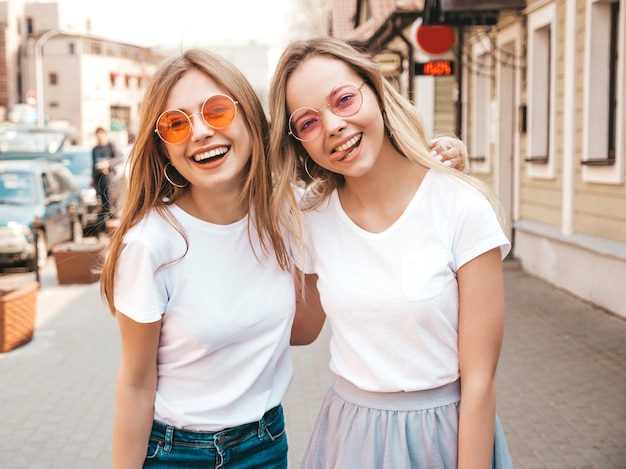 Un ritratto di due giovani belle ragazze sorridenti bionde dei pantaloni a vita bassa in vestiti bianchi della maglietta dell'estate d'avanguardia. . Modelli positivi che si divertono in occhiali da sole