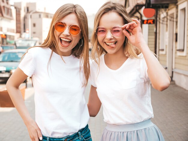 Un ritratto di due giovani belle ragazze sorridenti bionde dei pantaloni a vita bassa in vestiti bianchi della maglietta dell'estate d'avanguardia. . Modelli positivi che si divertono in occhiali da sole