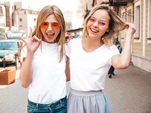 Un ritratto di due giovani belle ragazze sorridenti bionde dei pantaloni a vita bassa in vestiti bianchi della maglietta dell'estate d'avanguardia. . Modelli positivi che si divertono in occhiali da sole