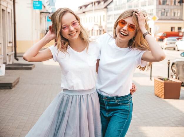 Un ritratto di due giovani belle ragazze sorridenti bionde dei pantaloni a vita bassa in vestiti bianchi della maglietta dell'estate d'avanguardia. . Modelli positivi che si divertono in occhiali da sole