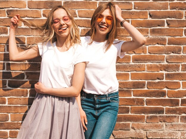 Un ritratto di due giovani belle ragazze sorridenti bionde dei pantaloni a vita bassa in vestiti bianchi della maglietta dell'estate d'avanguardia. Sexy spensierato. Modelli positivi che si divertono in occhiali da sole