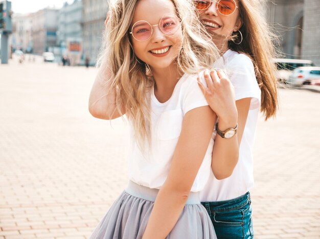 Un ritratto di due giovani belle ragazze sorridenti bionde dei pantaloni a vita bassa in vestiti bianchi della maglietta dell'estate d'avanguardia.