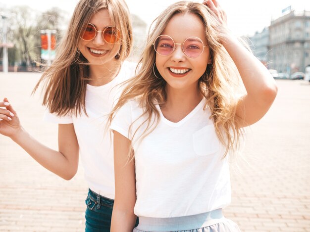 Un ritratto di due giovani belle ragazze sorridenti bionde dei pantaloni a vita bassa in vestiti bianchi della maglietta dell'estate d'avanguardia.