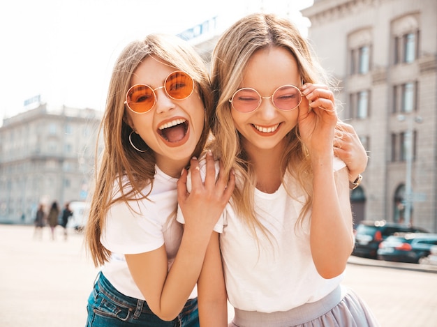 Un ritratto di due giovani belle ragazze sorridenti bionde dei pantaloni a vita bassa in vestiti bianchi della maglietta dell'estate d'avanguardia.