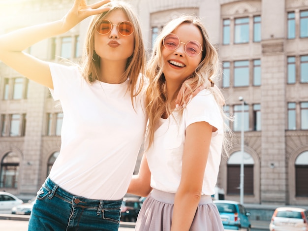 Un ritratto di due giovani belle ragazze sorridenti bionde dei pantaloni a vita bassa in vestiti bianchi della maglietta dell'estate d'avanguardia.