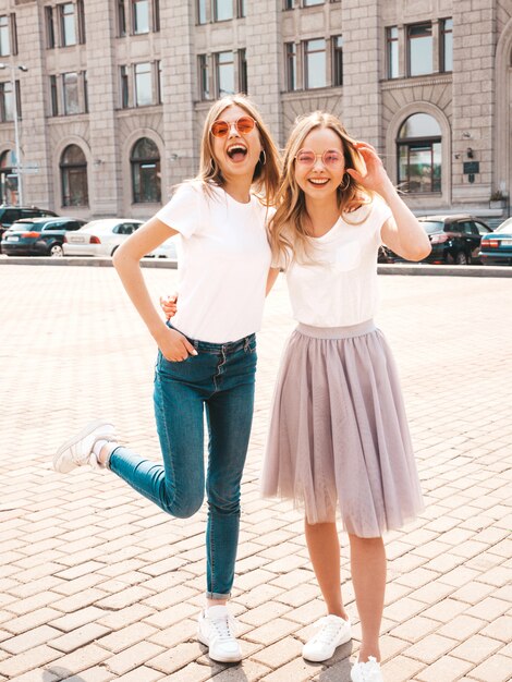 Un ritratto di due giovani belle ragazze sorridenti bionde dei pantaloni a vita bassa in vestiti bianchi della maglietta dell'estate d'avanguardia.