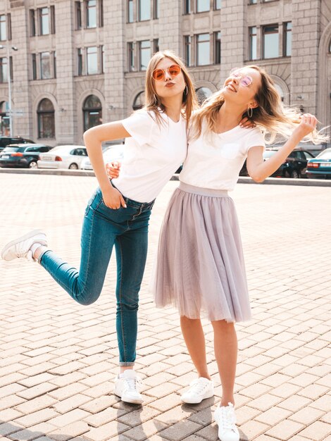 Un ritratto di due giovani belle ragazze sorridenti bionde dei pantaloni a vita bassa in vestiti bianchi della maglietta dell'estate d'avanguardia.